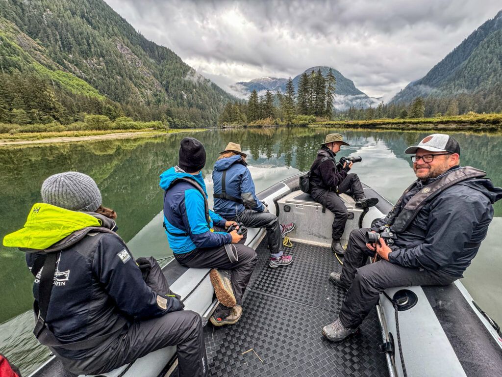 Five people riding in a motorboat on a calm mountain lake, surrounded by forested cliffs and cloudy skies
