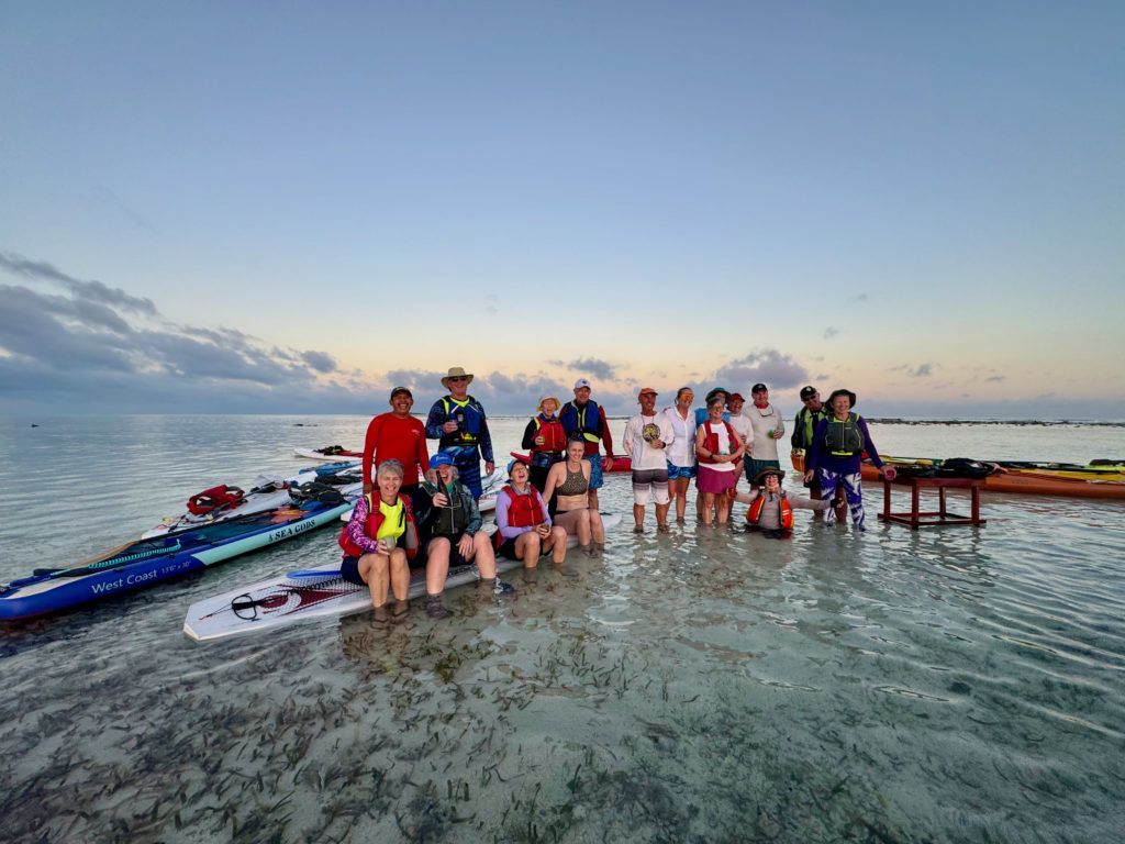 Group standing on paddleboards in shallow ocean water at sunset, with boards lined up nearby.