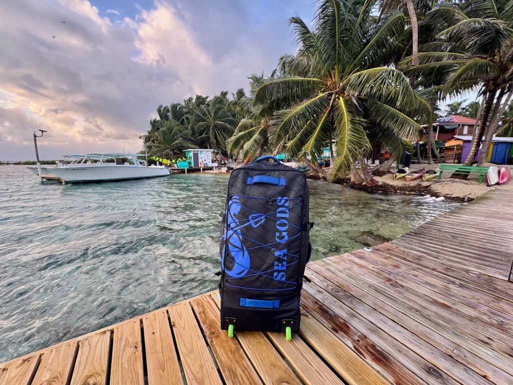 Blue wheeled backpack on a wooden dock beside turquoise water, boats, and palm trees at sunset