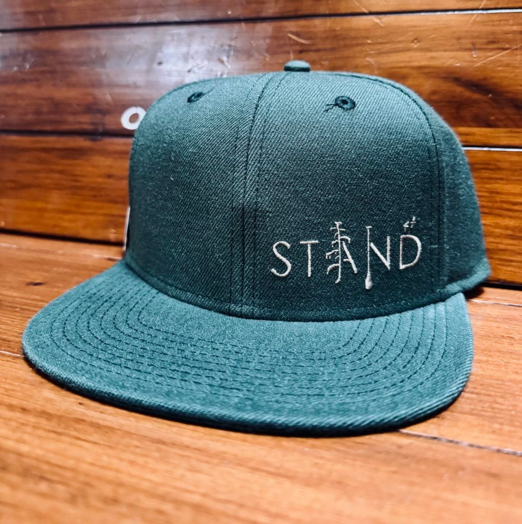 Teal flat-brim cap with “STAND” embroidered on the front, resting on a wooden surface
