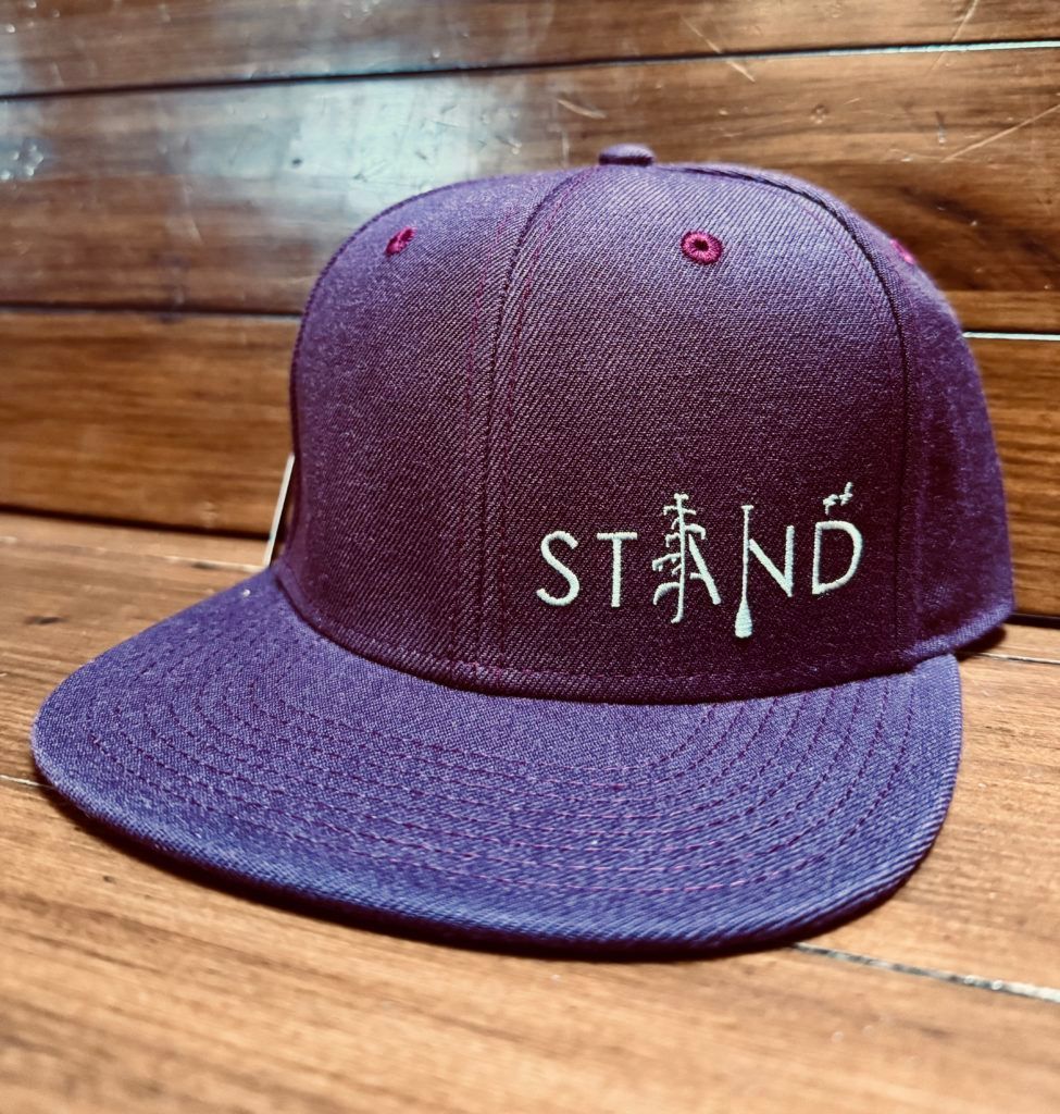 Purple STND baseball cap on a wooden surface, with white embroidered text and a small red button logo.