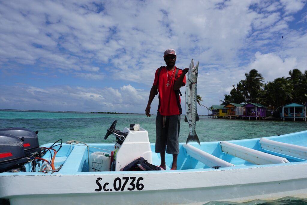 Man standing on a small boat in clear turquoise water near a tropical shore, holding a large fish.