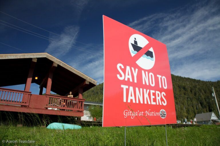 Red protest sign reading “SAY NO TO TANKERS” beside a house under a blue sky