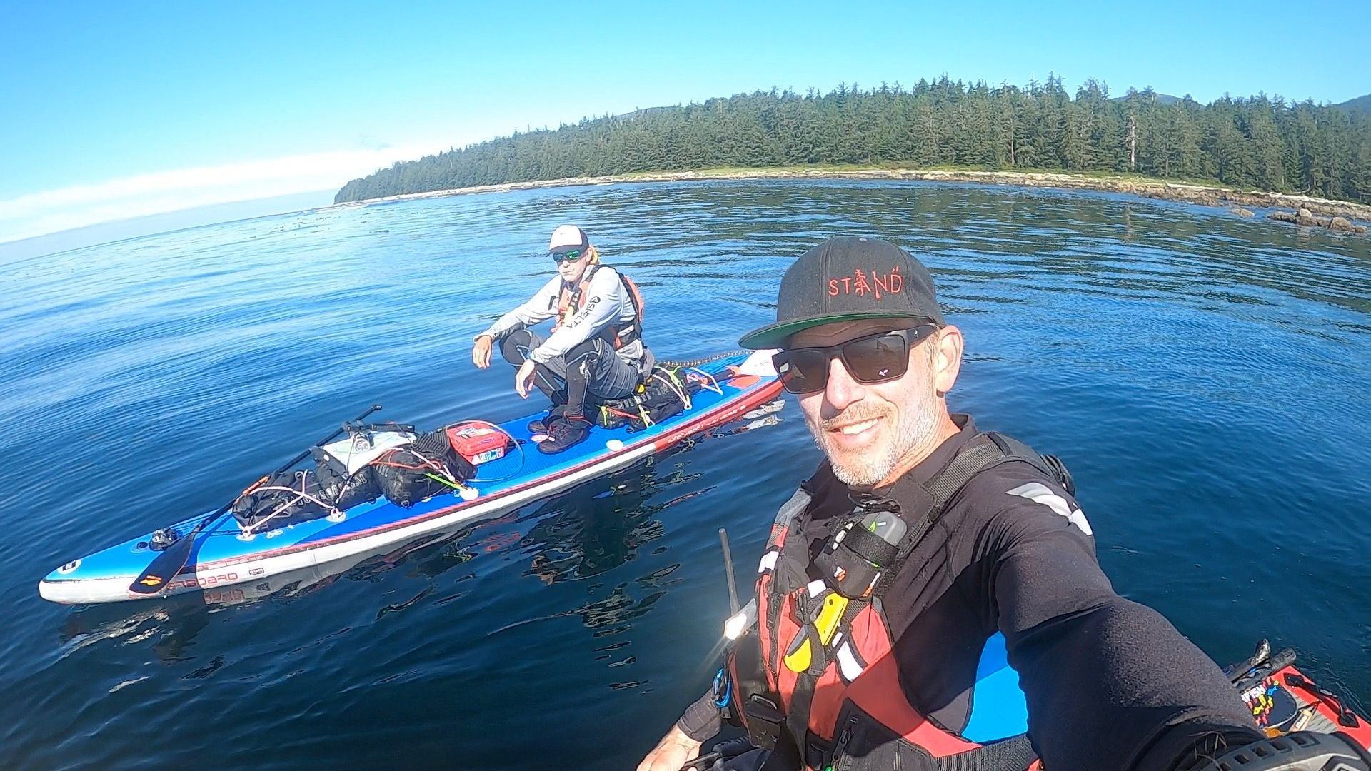Two kayakers on calm blue water near a forested shoreline, one taking a selfie in the foreground