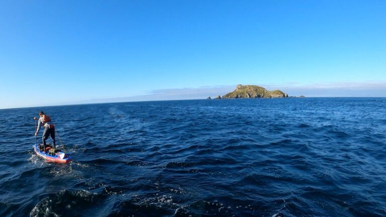 Person paddleboarding on blue ocean near a small rocky island under a clear sky