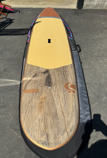 Top-down view of a paddleboard with a yellow deck pad and weathered wood-pattern finish on pavement