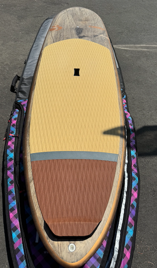 Top-down view of a paddleboard with a yellow deck pad and weathered wood-pattern finish on pavement