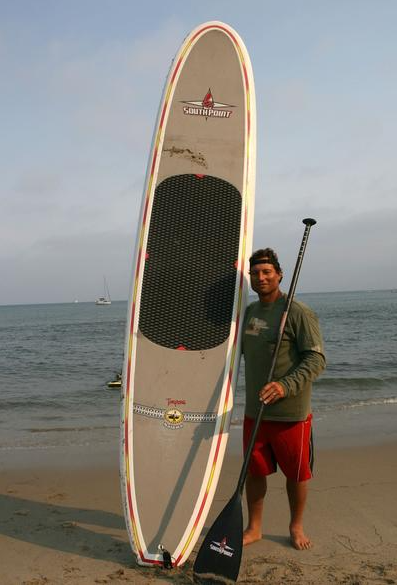 Man posing beside an upright paddleboard on a beach, holding a paddle with the ocean behind him.