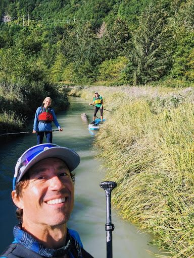Hikers paddling through a narrow river in a grassy canyon, with one smiling selfie in the foreground