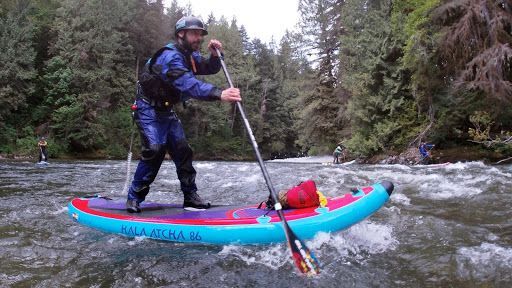Paddler standing on a blue inflatable raft, navigating a rocky river in a forested area
