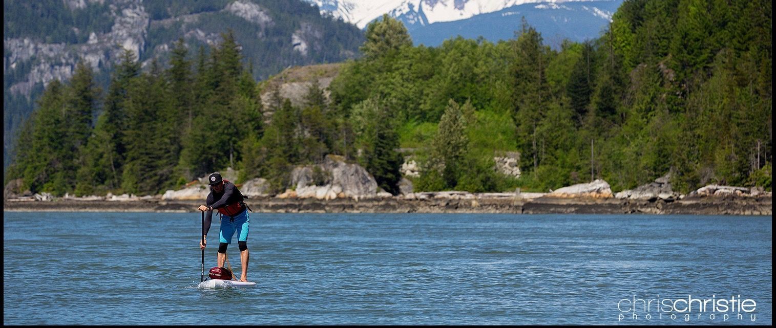 Two people paddleboarding on a blue river with forested mountains in the background