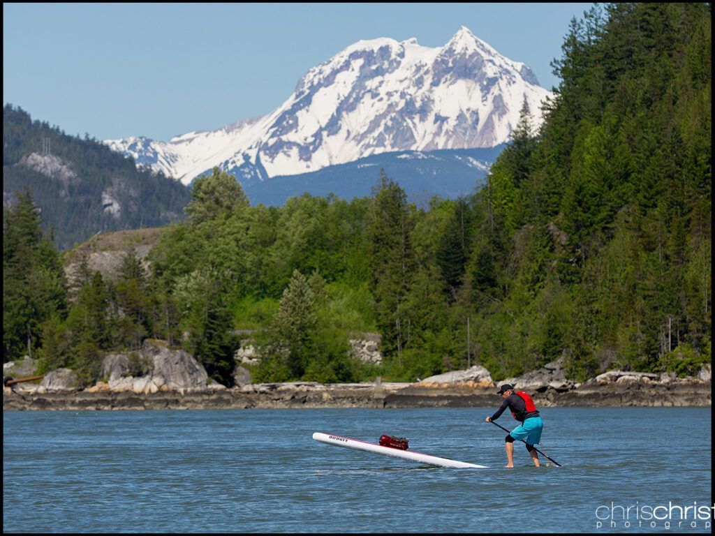 Person on a paddleboard in blue water with a snowy mountain and forested shoreline in the background