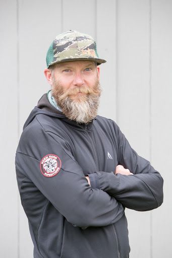 Man in a camo cap and dark jacket stands with arms crossed against a gray wall.