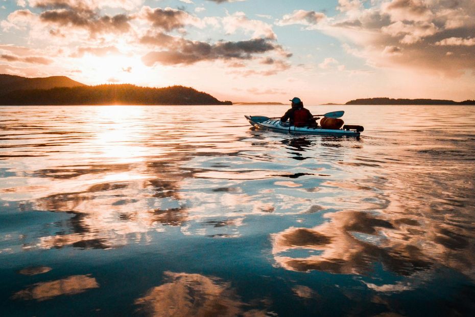 Person kayaking on calm water at sunset, with golden reflections and distant shoreline