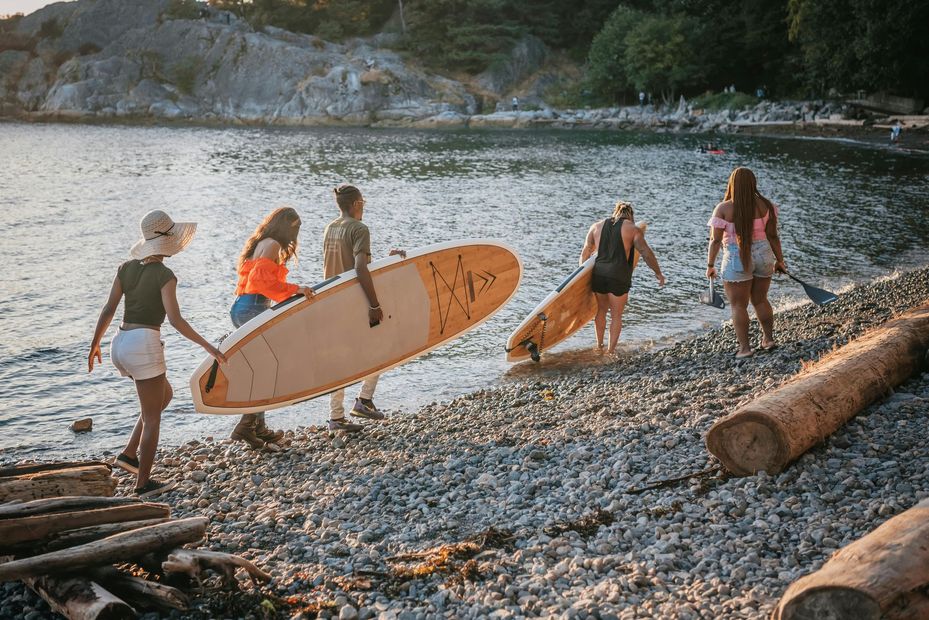 People carrying paddleboards and walking along a rocky beach by the water