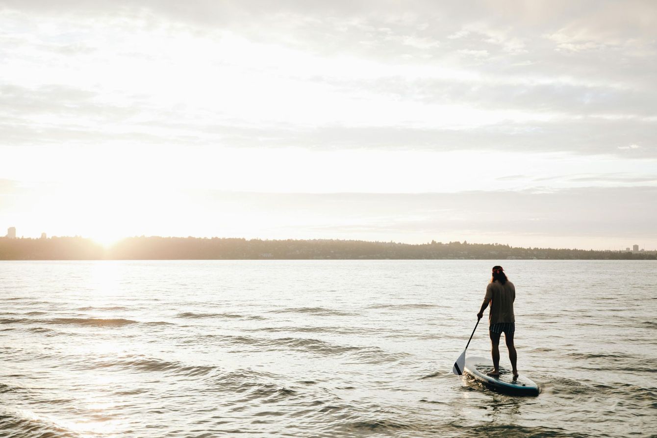 Person paddleboarding on calm water at sunrise, with a bright horizon and distant shoreline
