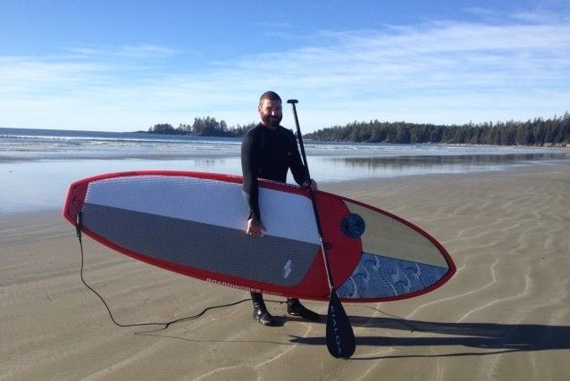 Person standing on a sandy beach holding a red and gray paddleboard with a paddle beside it.