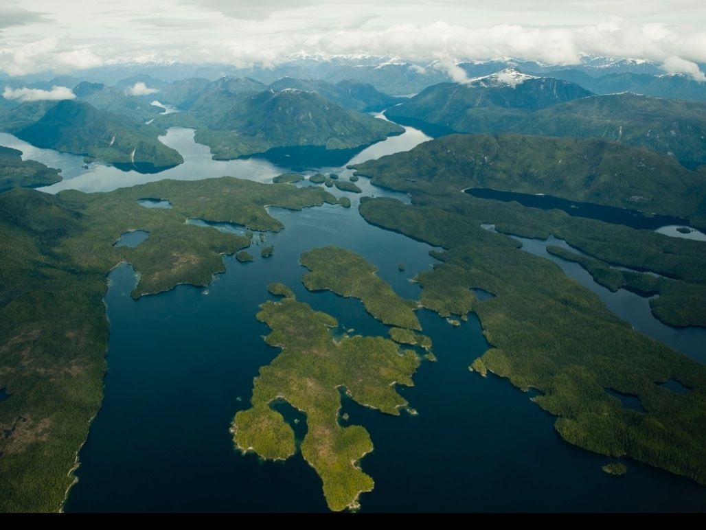 Aerial view of forested islands and winding lakes beneath cloudy mountains.
