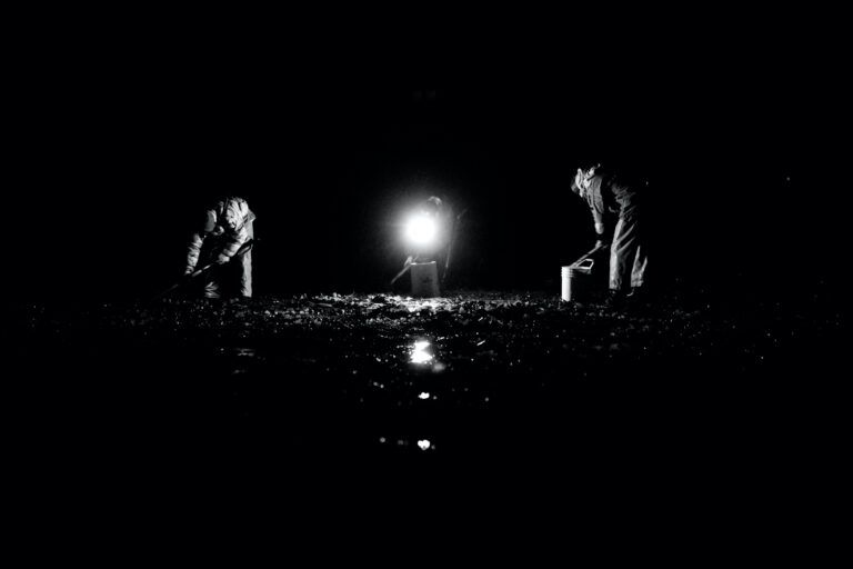 Two people crouch in a dark field lit by a bright central light at night.