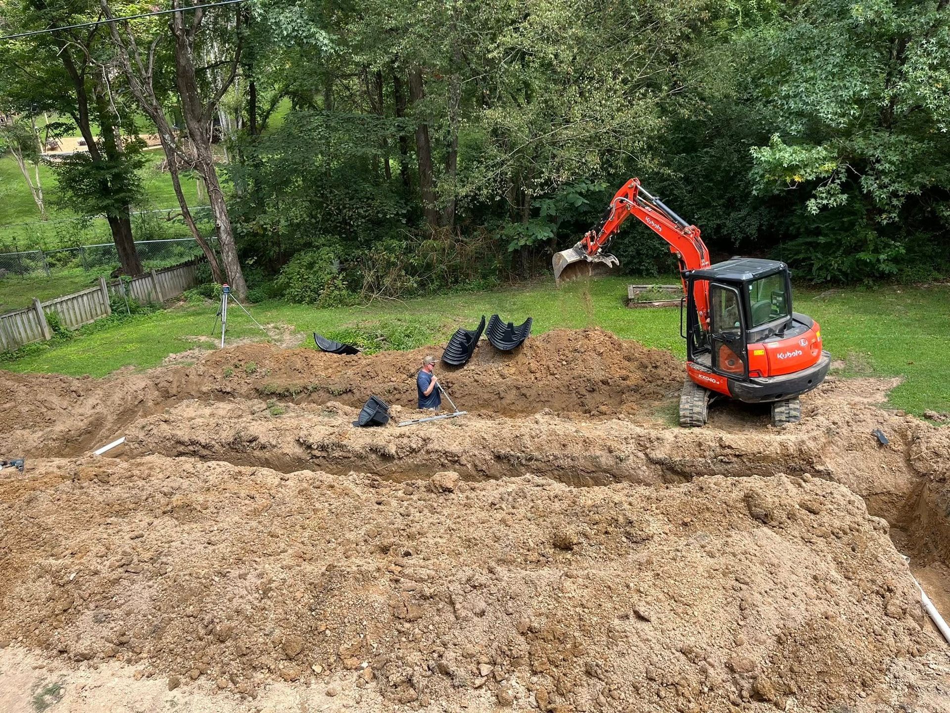 Orange excavator digging a muddy lot with workers nearby, surrounded by trees