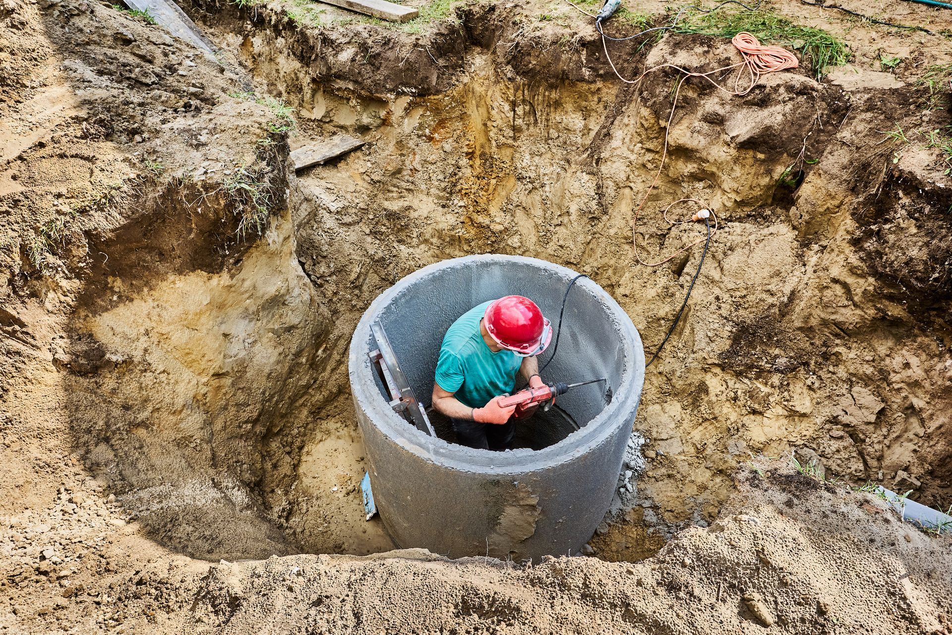 Worker in a concrete well shaft, crouching inside a circular pipe amid an excavation site.