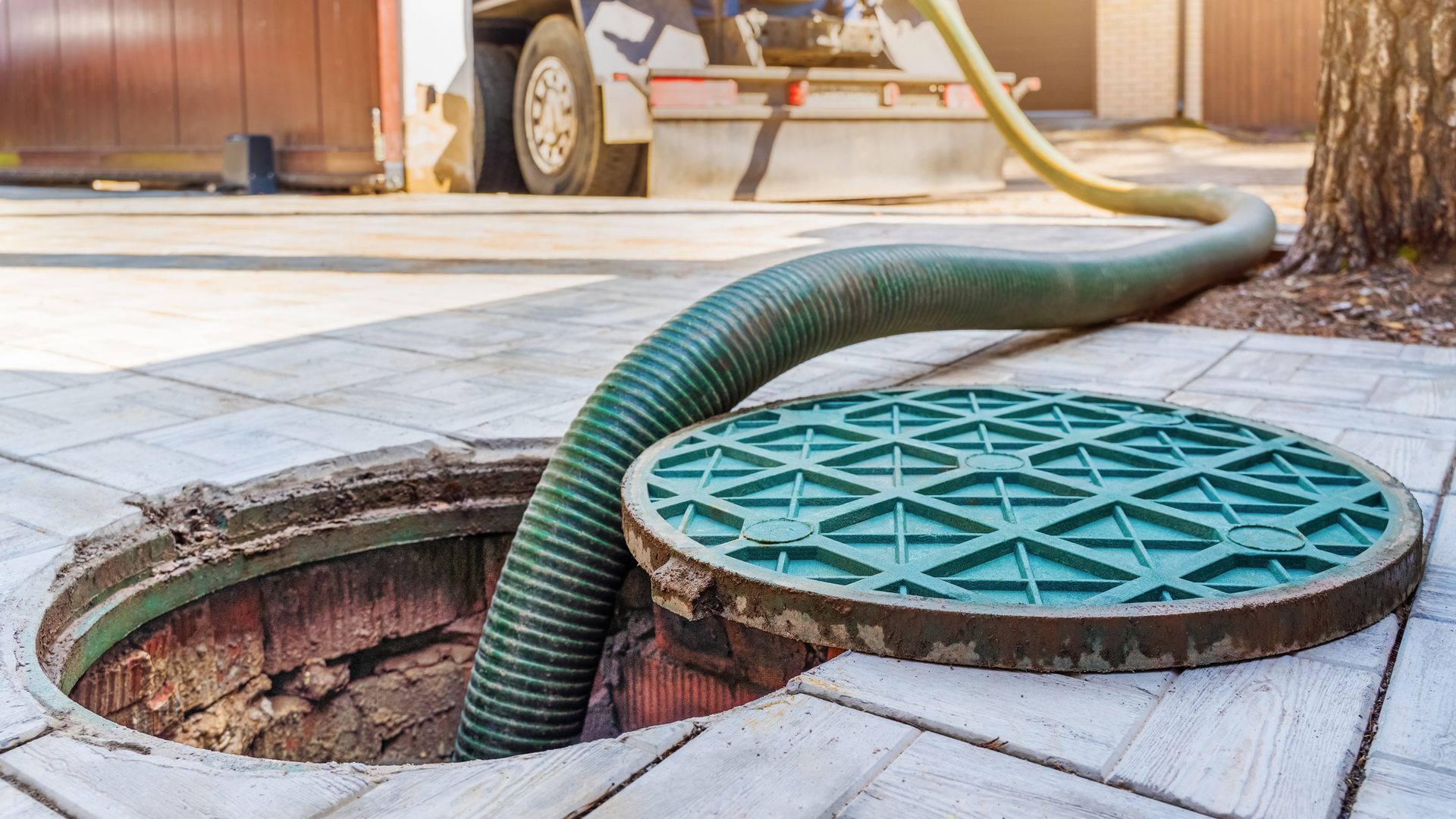 Green hose entering open manhole on a wooden deck, with a round teal cover nearby.