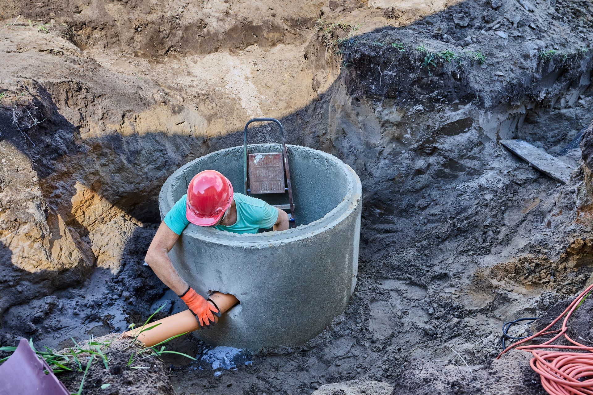 Worker in red hard hat and teal shirt inside a concrete well, digging in muddy trench.