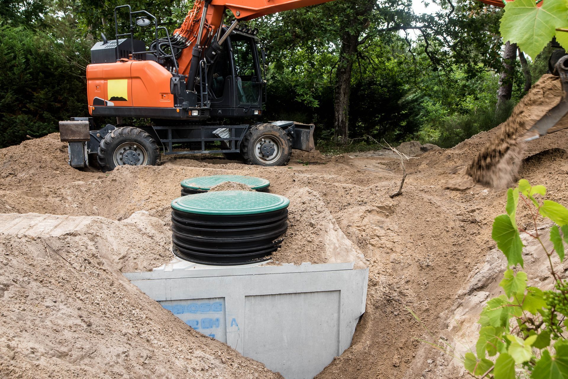 Orange excavator at a dirt construction site beside a concrete utility chamber with a green cover.