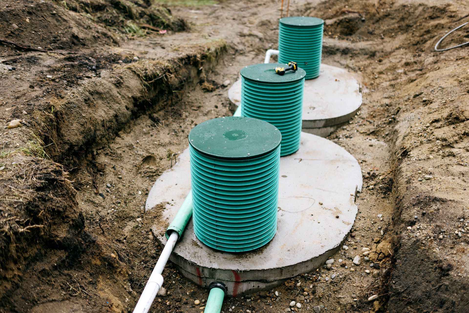 Two green septic tanks installed in a dirt excavation with white lids and exposed pipes