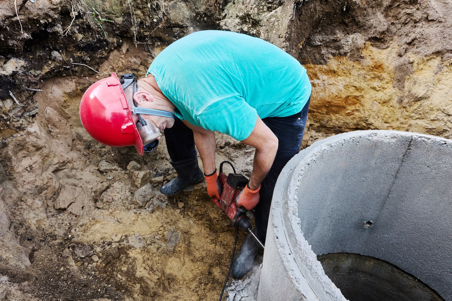 Worker in a red hard hat and teal shirt crouches in a muddy trench beside a concrete pipe.