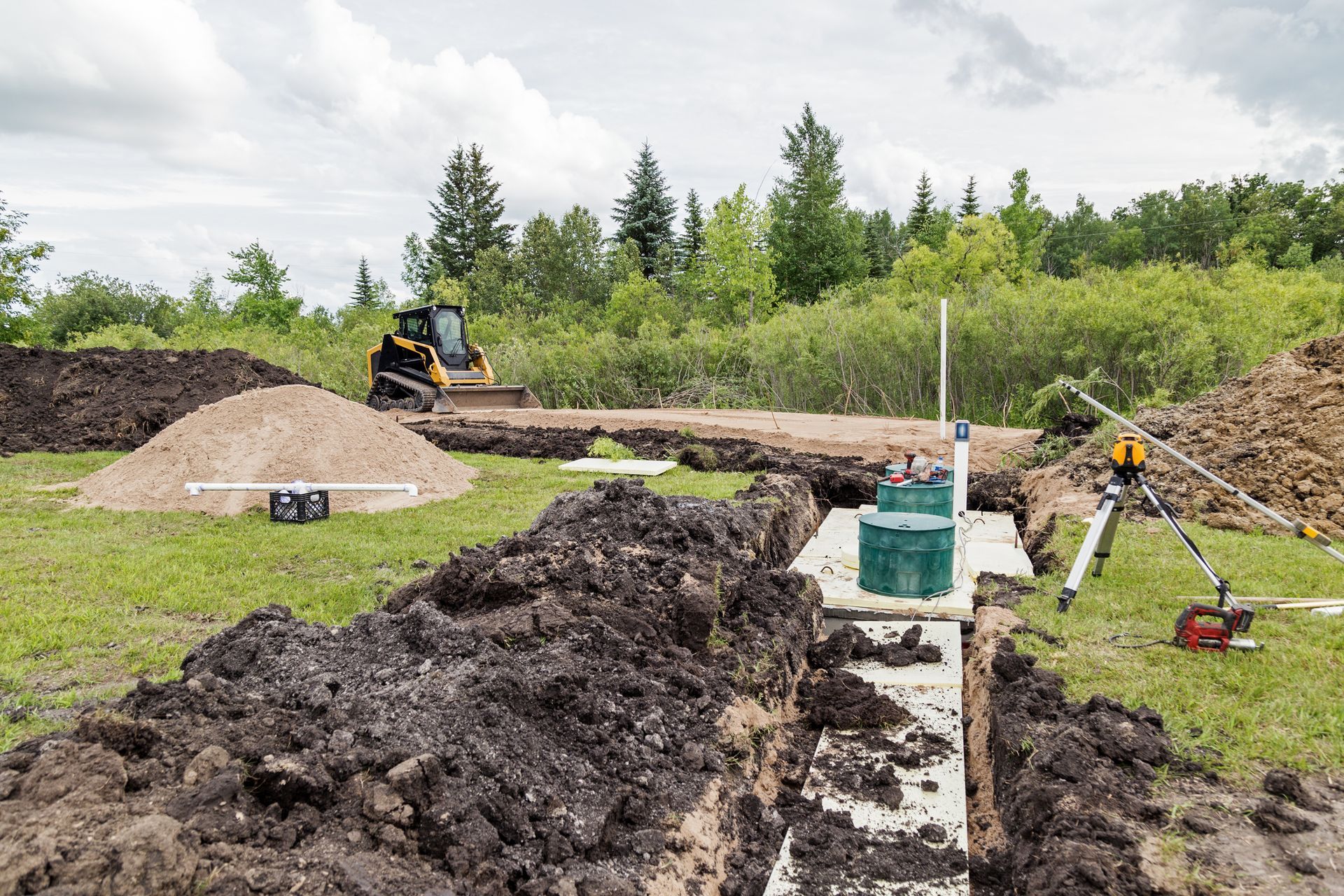 Construction site with trenches, excavator, survey tripod, and utility equipment on grassy field