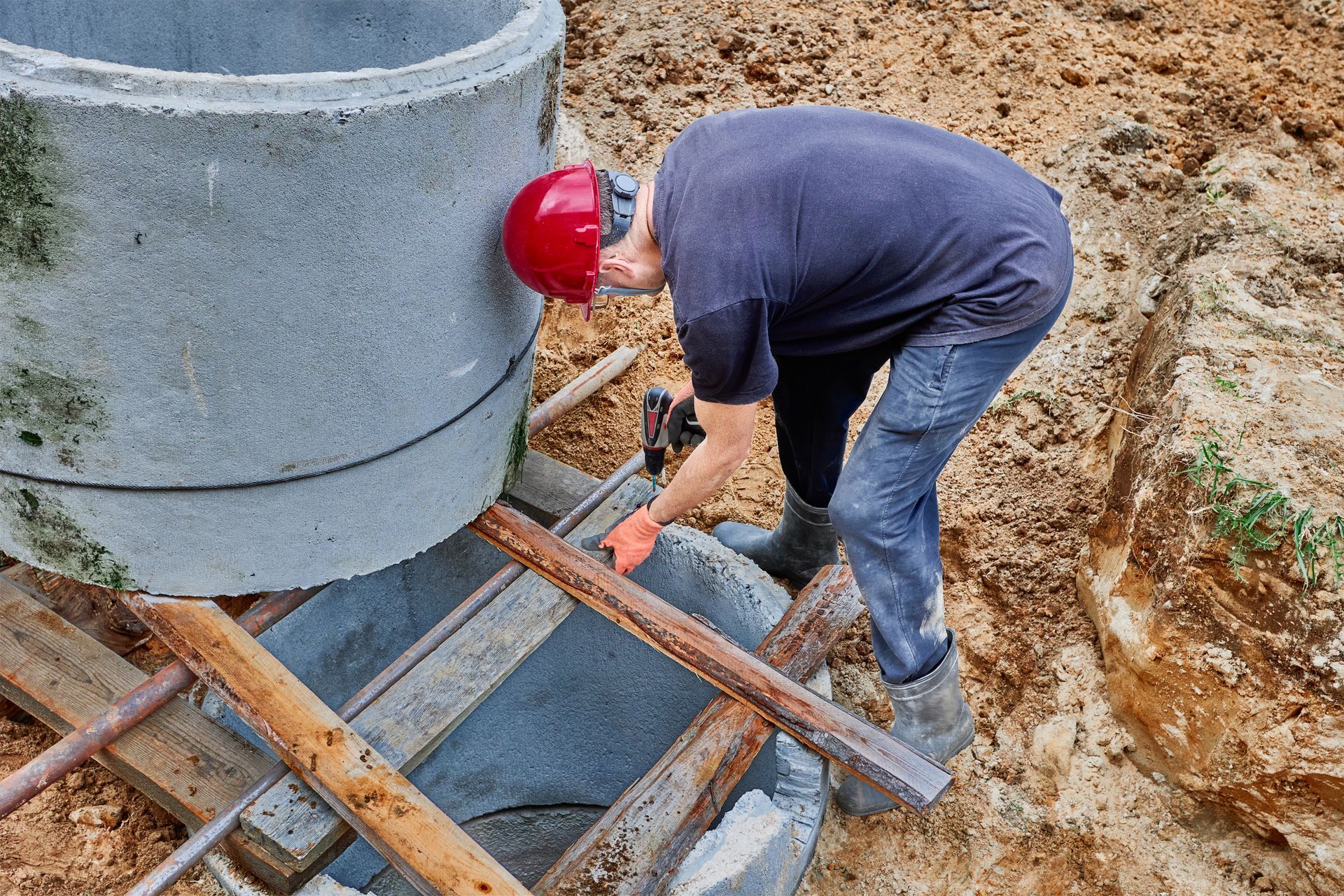 Worker in red hard hat bending over concrete formwork at a muddy construction site