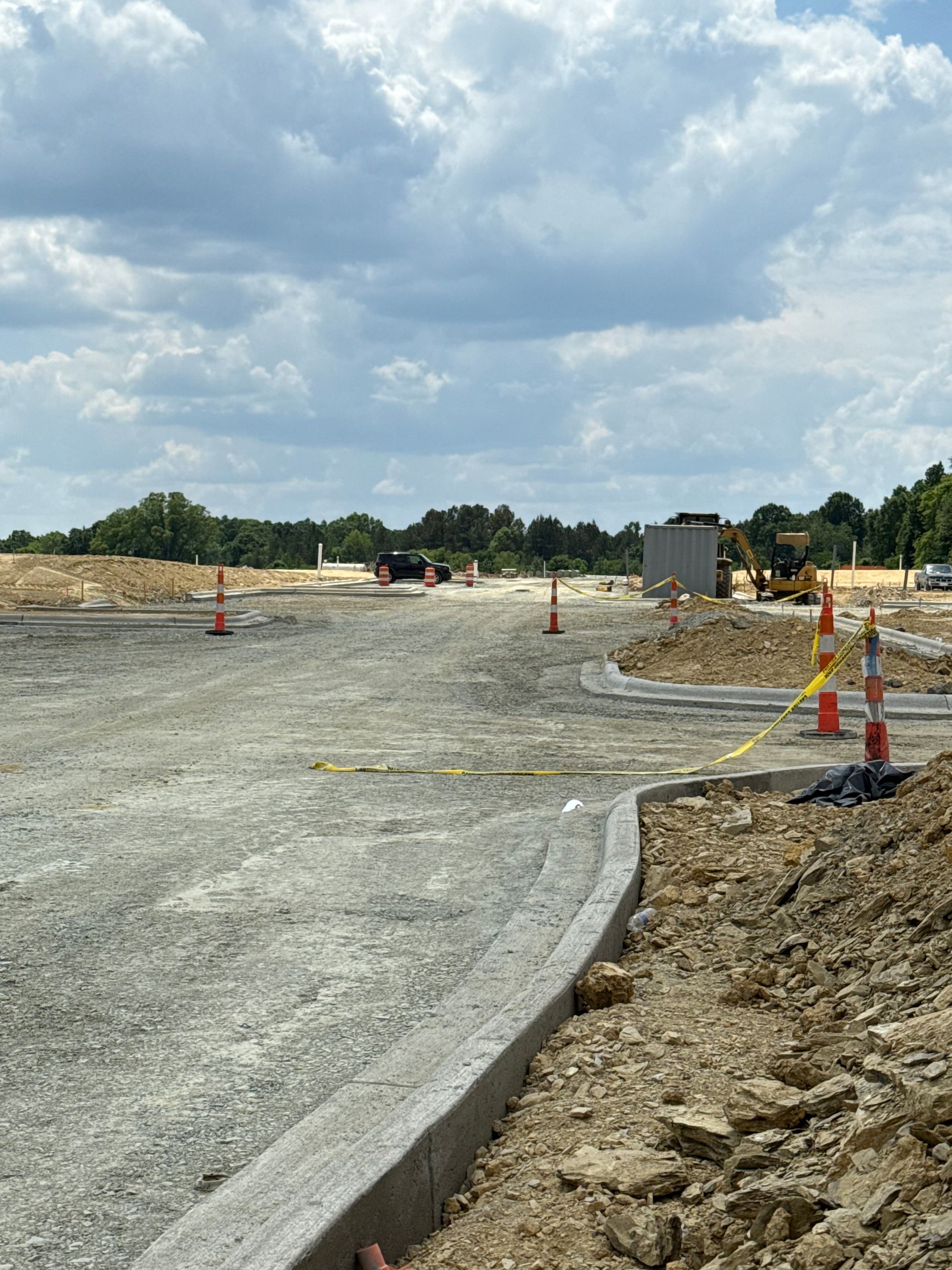 Road construction site with gravel, orange cones, and concrete curb under a cloudy sky
