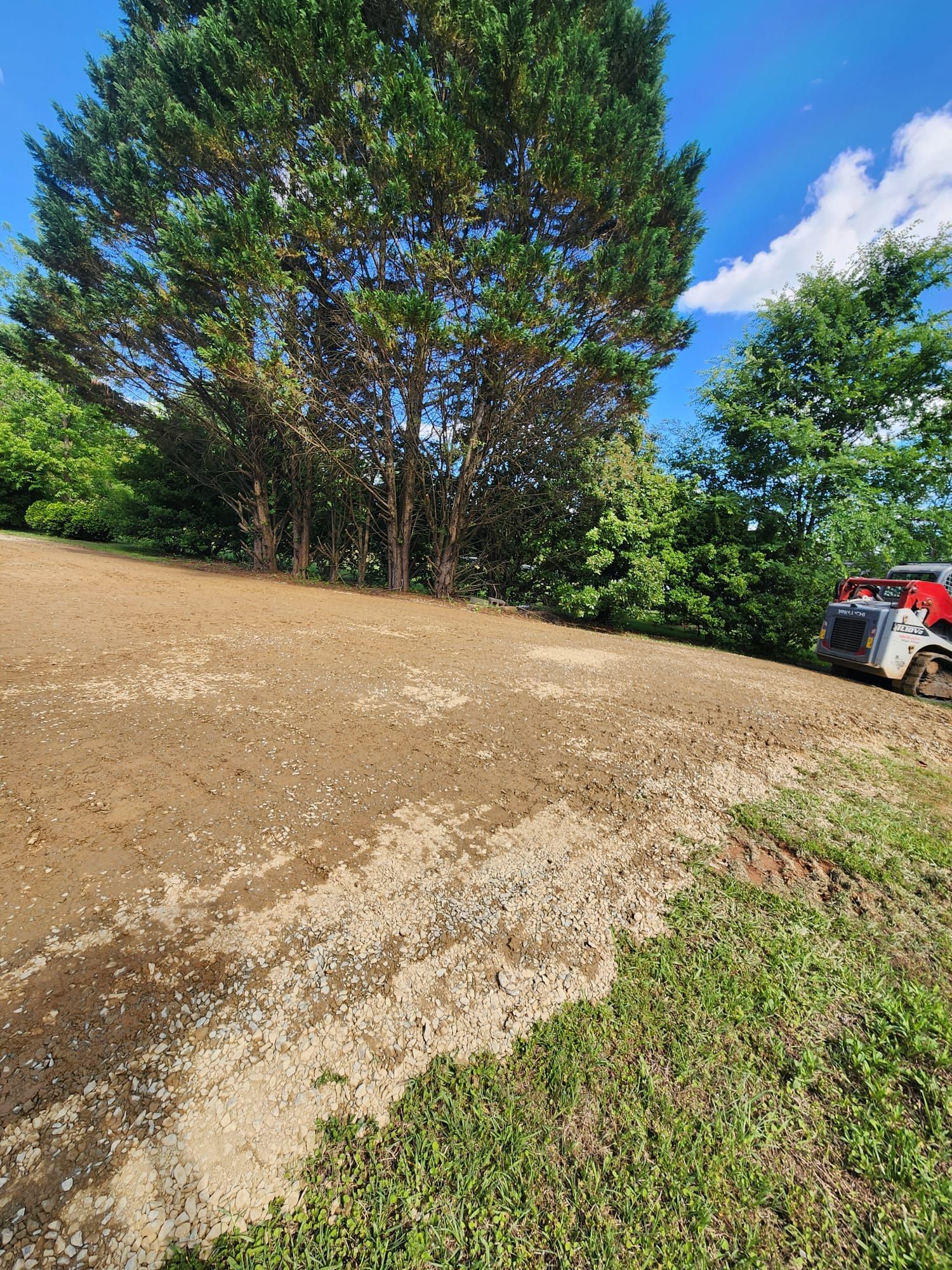 Gravel driveway curving past tall trees and green grass, with part of a red off-road vehicle at right.