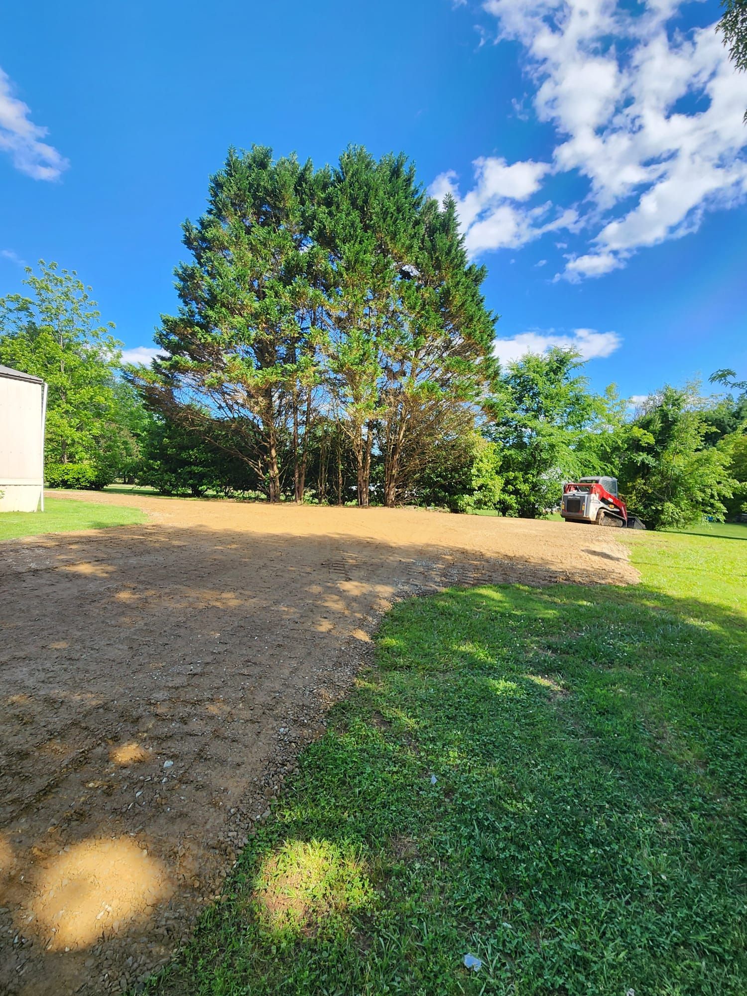 Gravel driveway beside a grassy yard with tall trees under a bright blue sky and clouds