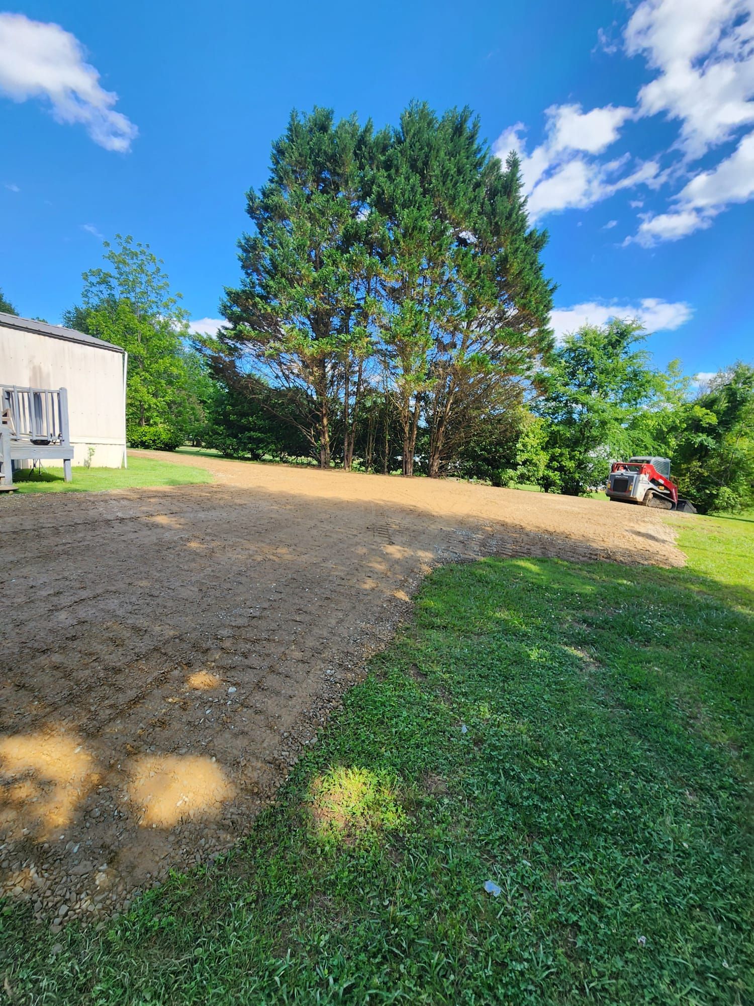 Gravel driveway beside a white building, with a large tree and red tractor under a bright blue sky.