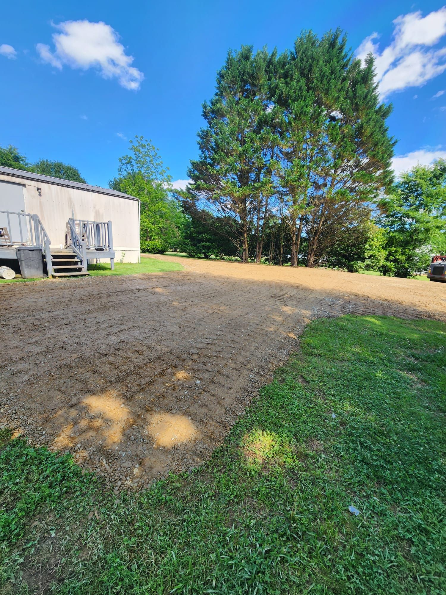 Gravel driveway beside a white building, with a large tree and green lawn under a blue sky.
