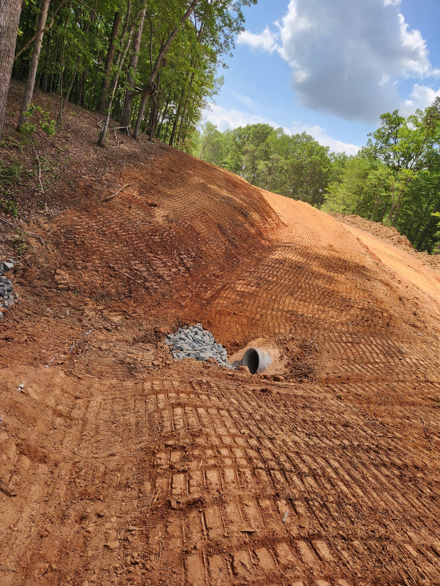 Dirt road embankment with a drainage pipe and rutted mud under a partly cloudy sky, surrounded by trees