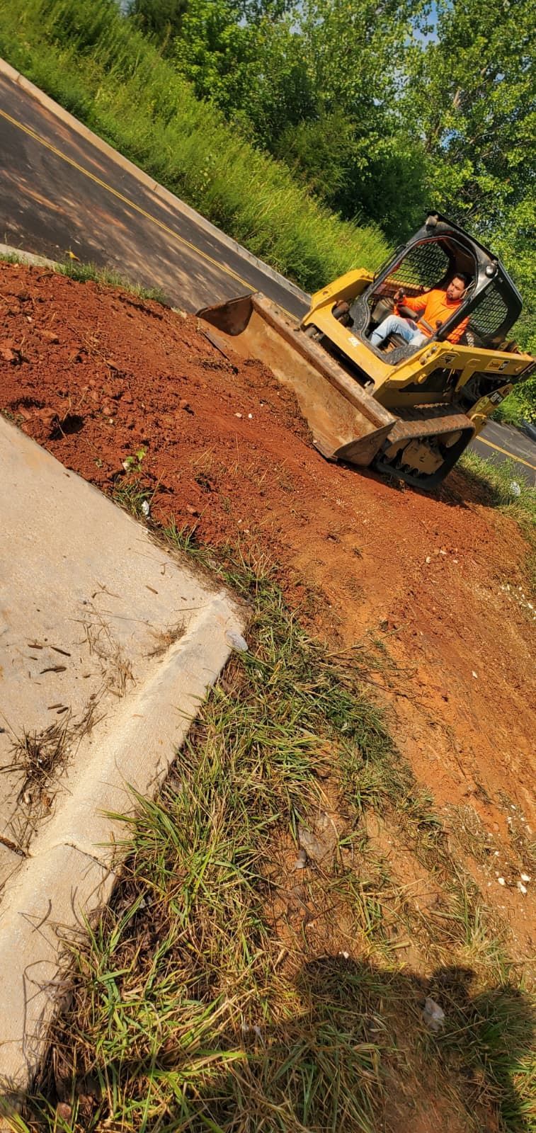 Yellow backhoe moving red dirt beside a curb on a roadside slope