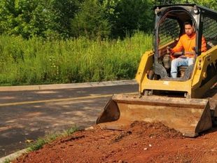 Yellow skid steer pushing red dirt beside a paved road with trees in the background
