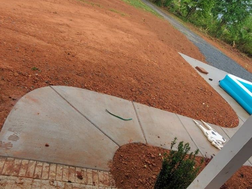 Concrete path beside red dirt slope and curb, with a blue pipe and small shrub.