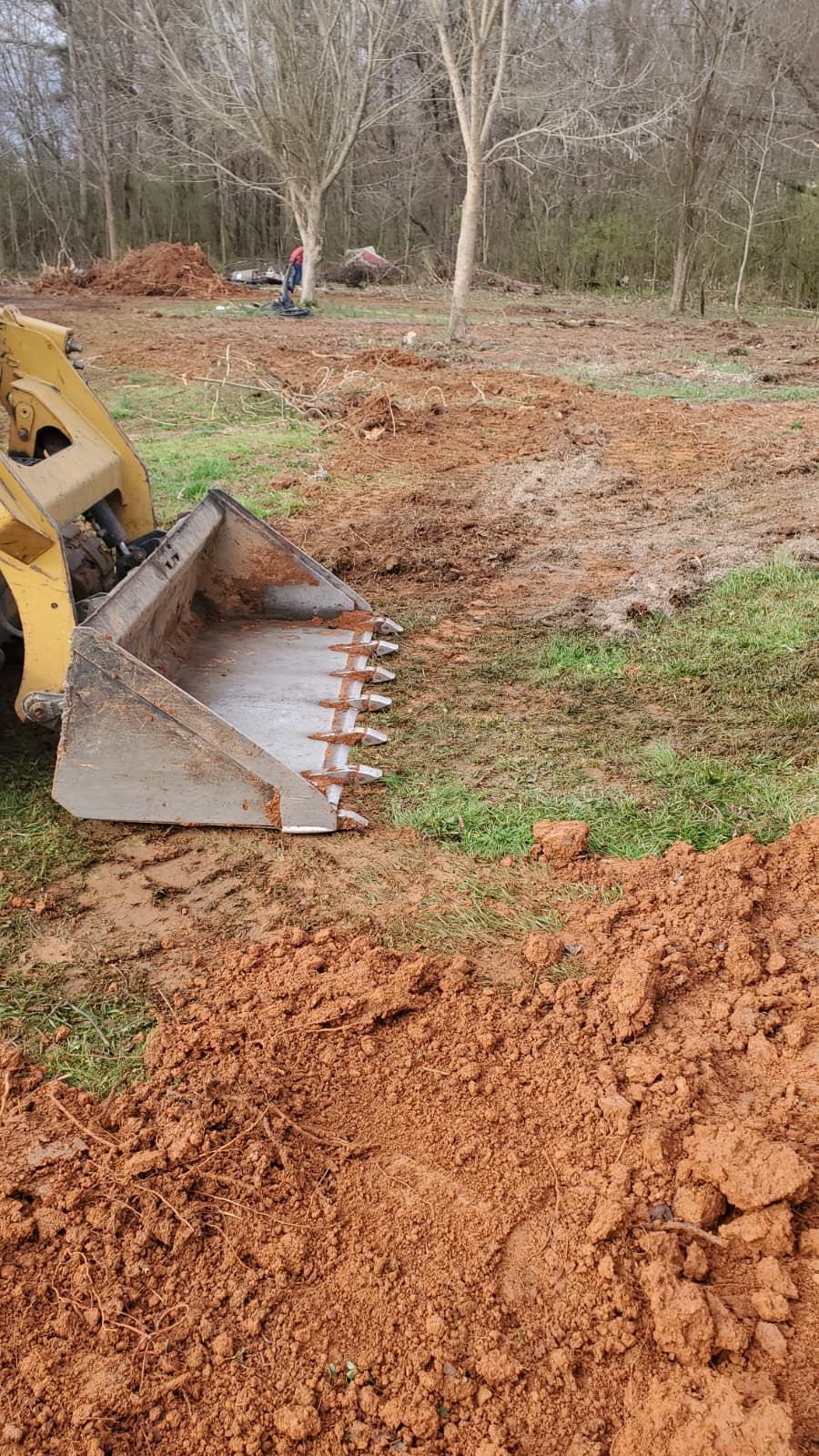 Bulldozer blade pushing red dirt across a muddy construction site with bare trees nearby