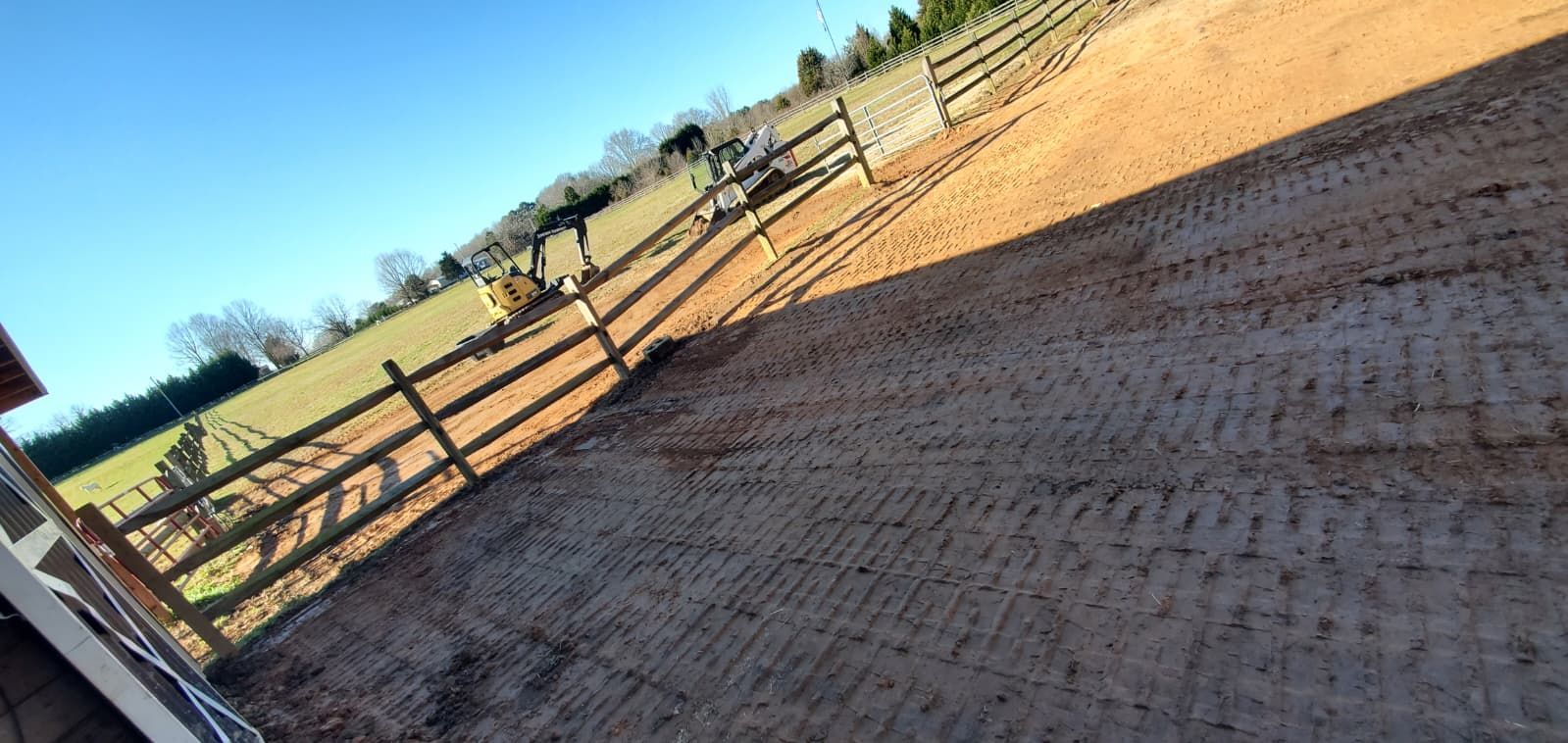 Tilted view of a fenced dirt paddock beside green fields under a blue sky