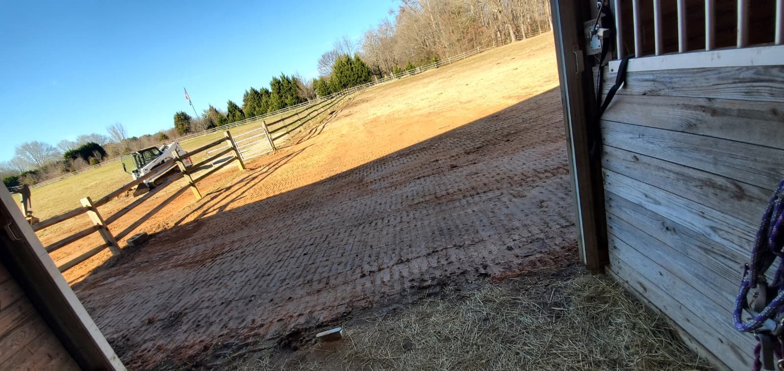 Tilted view of a fenced dirt yard beside a wooden barn under a clear blue sky