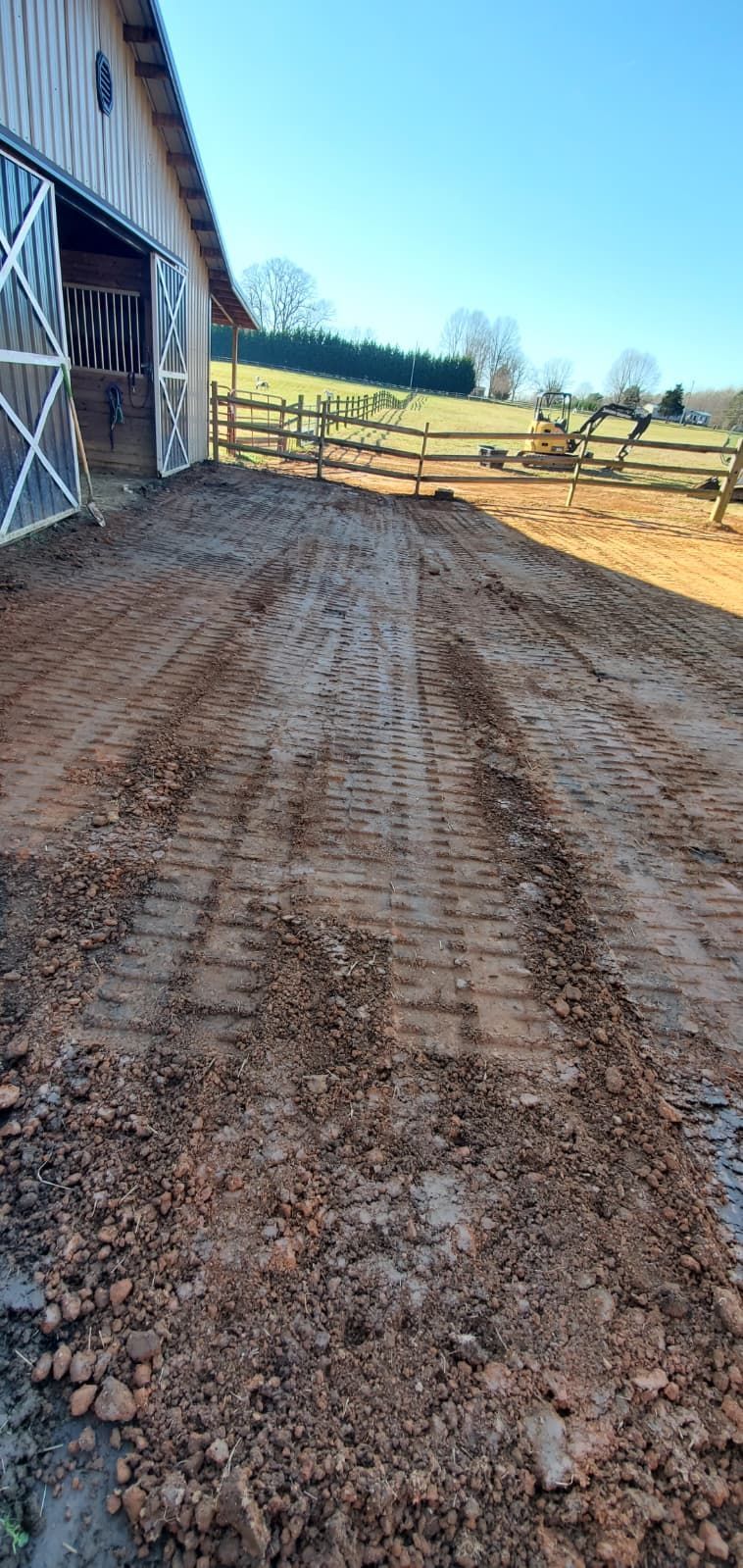 Muddy dirt lane beside a barn, leading to a fenced pasture under a clear blue sky.