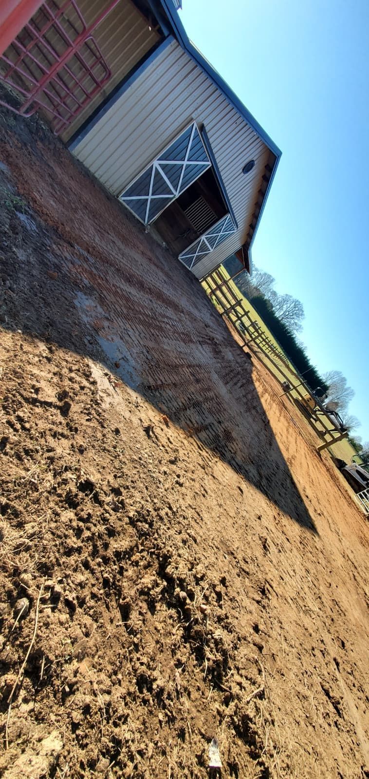 Tilted view of a brick barn with open white doors beside a bare dirt field under a blue sky