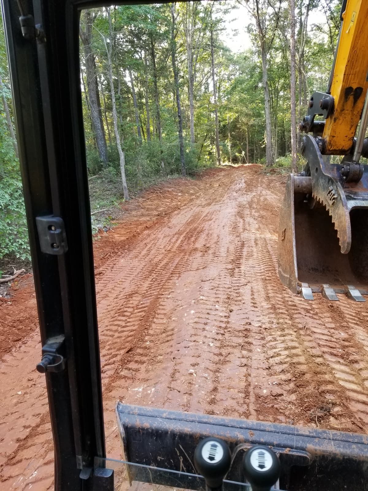 View from a cab of a yellow excavator grading a dirt road through a wooded area