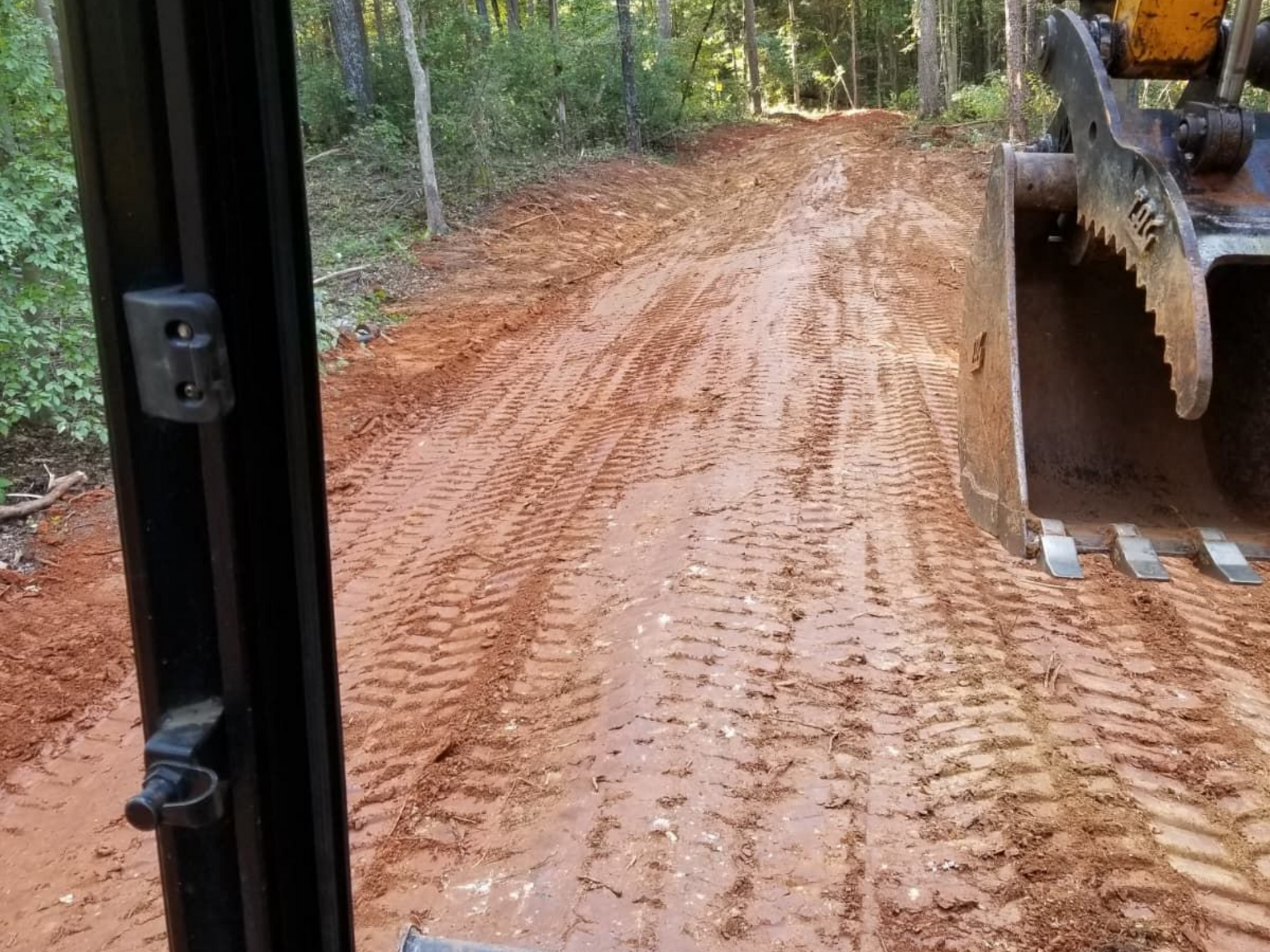 Inside a construction vehicle, a muddy dirt road with tire tracks and a backhoe bucket on the right.