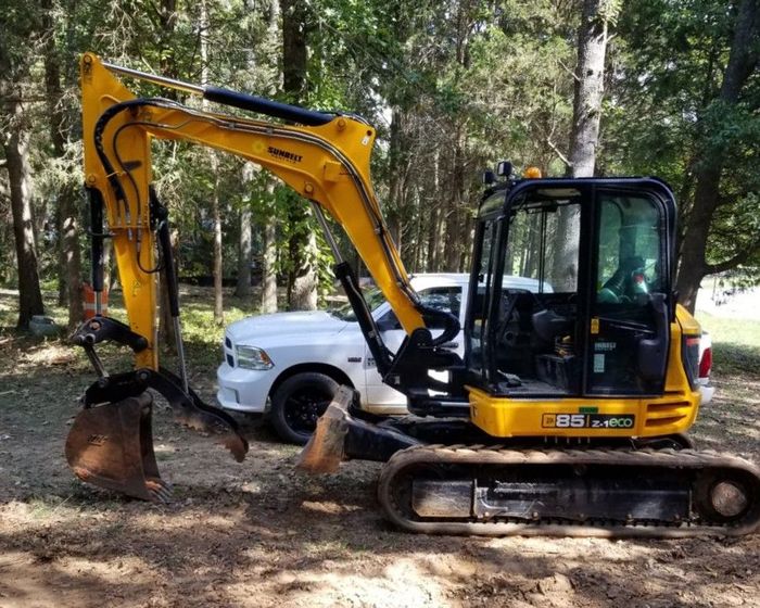 Yellow excavator beside a white pickup truck in a wooded outdoor area