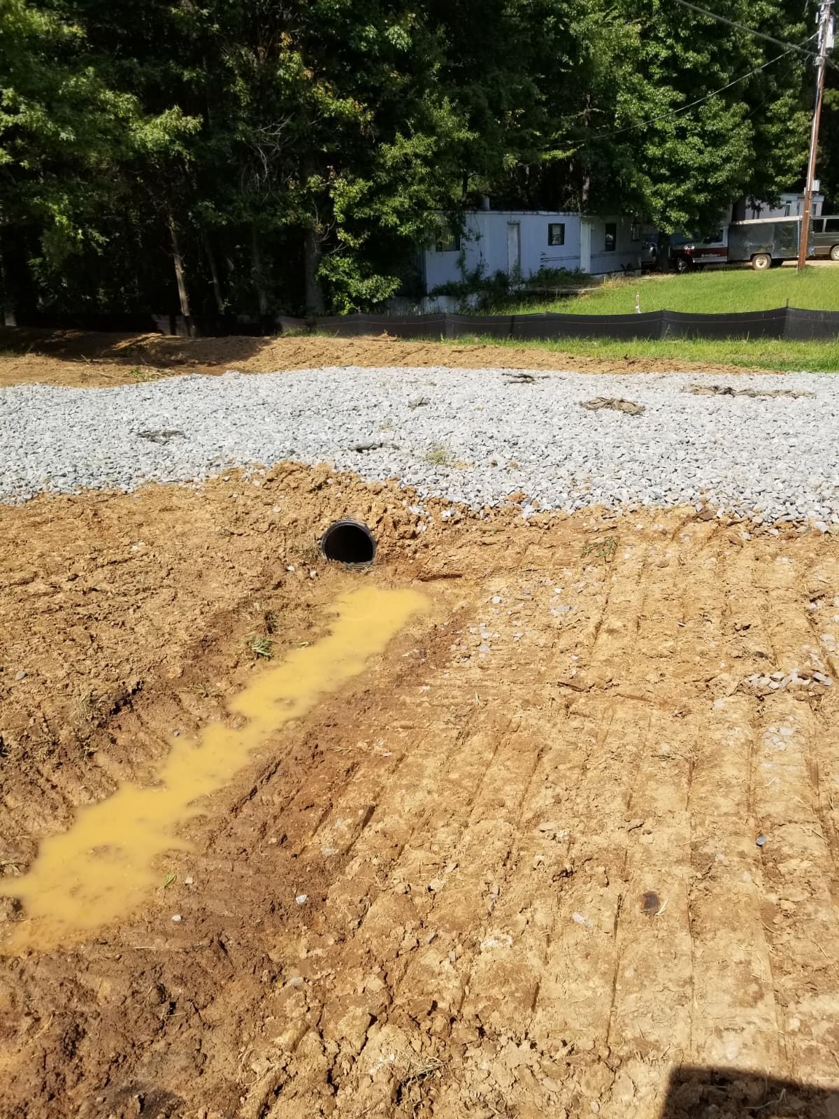 Gravel road under construction with muddy dirt, a drainage pipe, and trees in the background